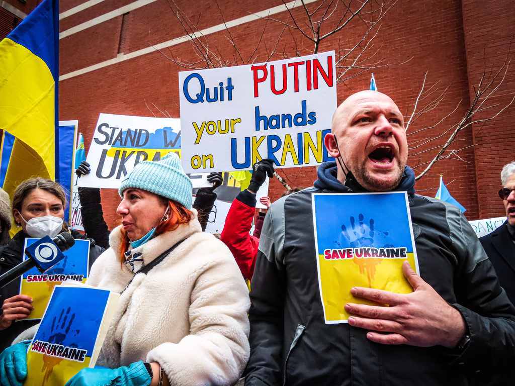 Protestantes contra a invasão da Rússia, segurando placas com as cores da bandeira da Ucrânia, escrito "Save Ukraine", "Putin, tire as mãos da Ucrânia", "Apoiamos a Ucrânia" etc